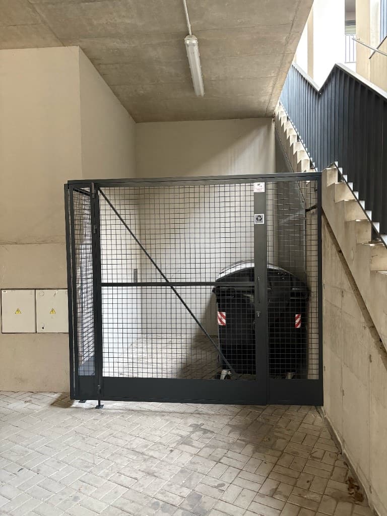 Ocelová síťová klec na odpadové kontejnery s dvoukřídlou bránou pod schodištěm bytového domu, v tmavě šedé